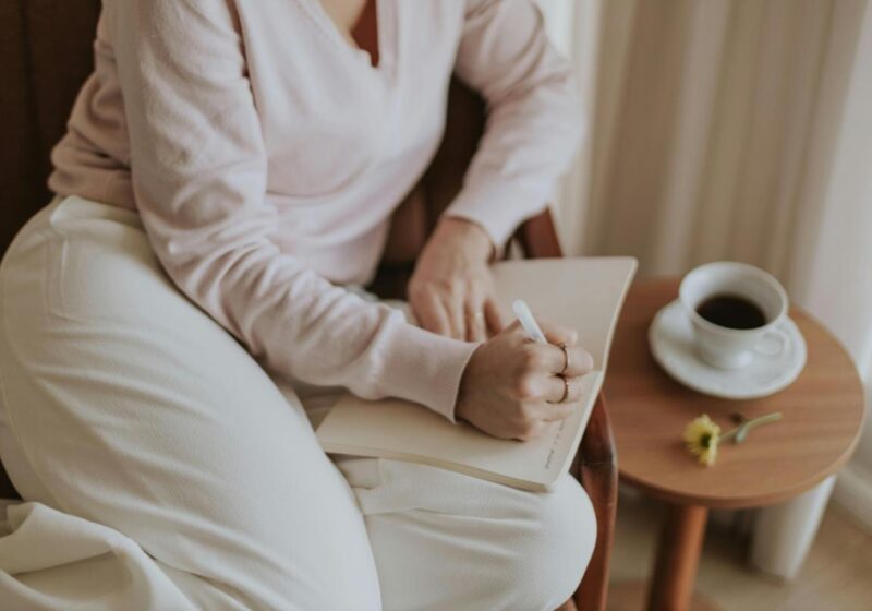 A woman in a soft beige sweater journaling in a notebook while sitting at a wooden table with a cup of coffee, illustrating a soft morning routine for a calm start.